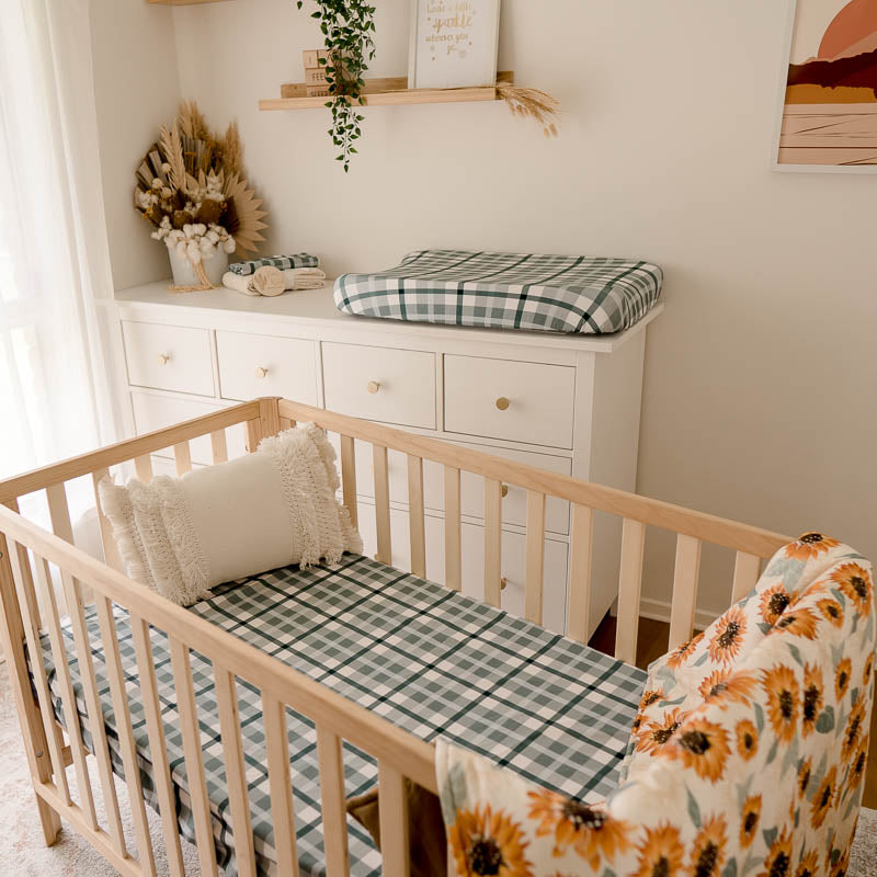 Full set of blue plaid items from snuggly jacks placed in a nursery with a sunflowers cot quilt hanging over the end of a pine cot.