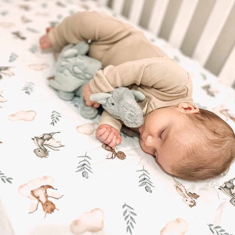 The cutest of babies sleeping while snuggling a plush toy in a cot. The sheet is white with little dragons, green leaves and soft brown clouds all over it. 