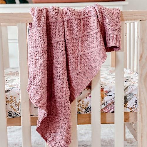 A warm and luxurious cotton knitted blanket in rose pink color from Snuggly Jacks, a premium Australian brand of knitted blankets and throws, draped over the side of a pine cot.
