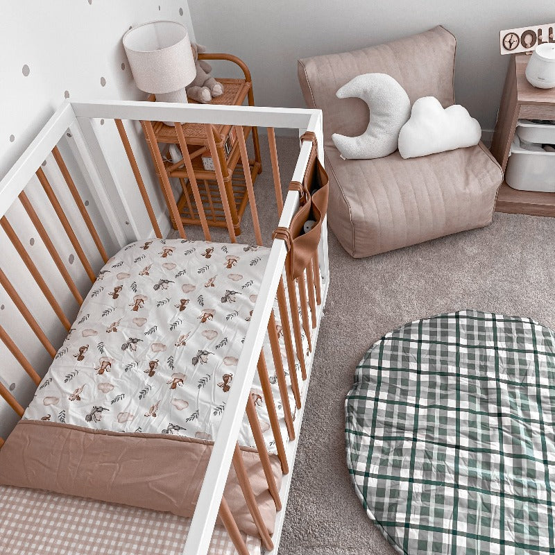 Modern nursery set with a blue plaid cotton playmat, soft chair in one corner, a rattan shelf and a pine cot in the bottom corner made up with the a cotton fitted sheet and a cotton cot quilt.