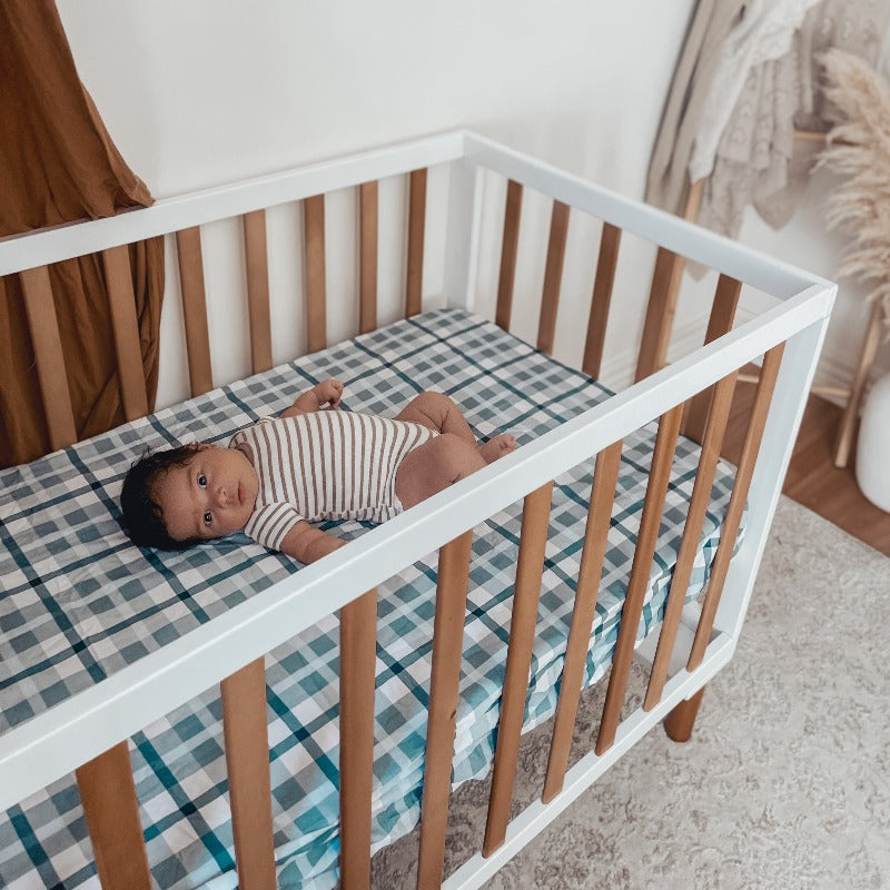 A Pine cot with white accents housing a baby loking at the camera while relaxing on a plaid patterned cotton cot sheet