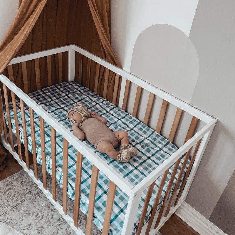 Sweet little baby wearing booties and a knitted beanie laying on a blue plaid cot sheet in a pine cot