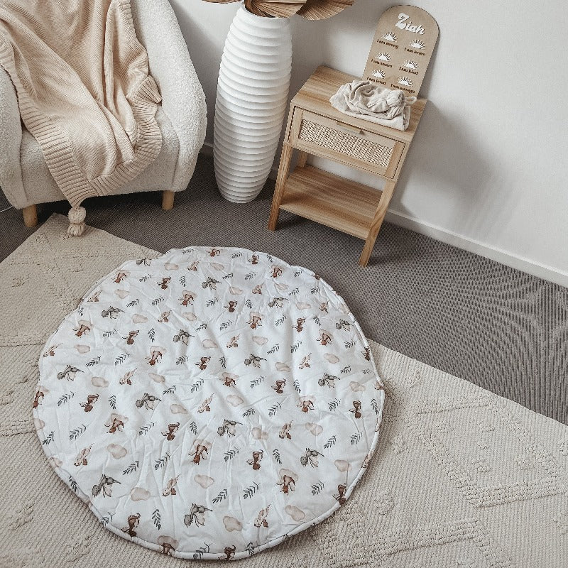 A 100% cotton playmat with adorable dragons laid out on the floor of a modern nursery with and arm chair in one corner and a pine draw with rattan details