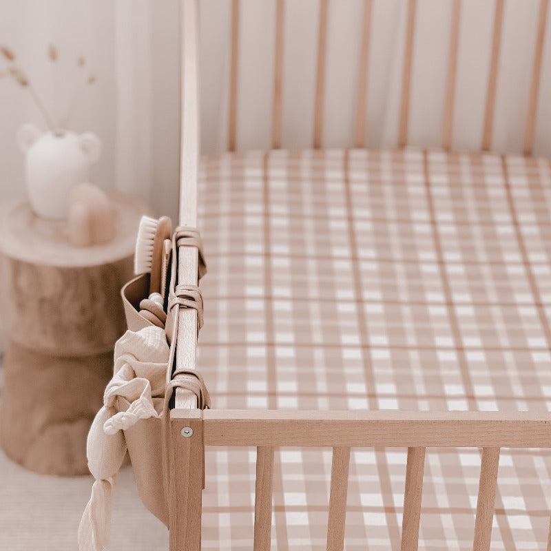Close up of a wooden cot with a nursery organiser hanging on one side a wooden bedside table in the back ground and a brown and white plaid fitted cot sheet
