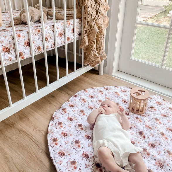 Baby laying on a floral play mat in a nursery with a cot, plush bunny and wooden toy