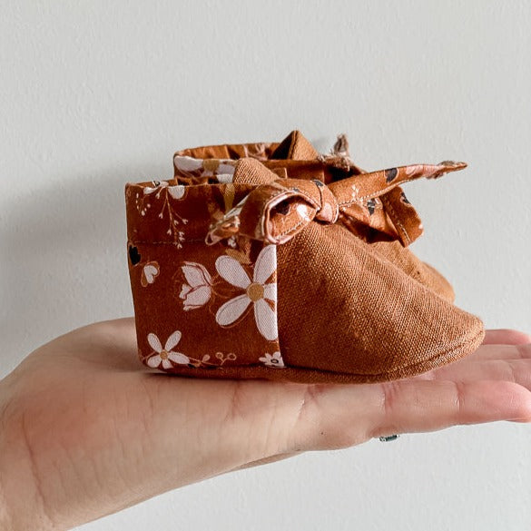 a Pair of Brown Linen baby booties with floral backing on a womens hand against a white wall