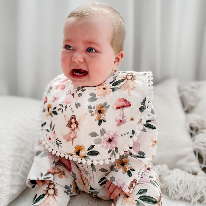 Baby sitting while wearing a Fairy Garden print bib, featuring enchanting fairies, vibrant florals, and absorbent microfiber backing for mess-free feeding.