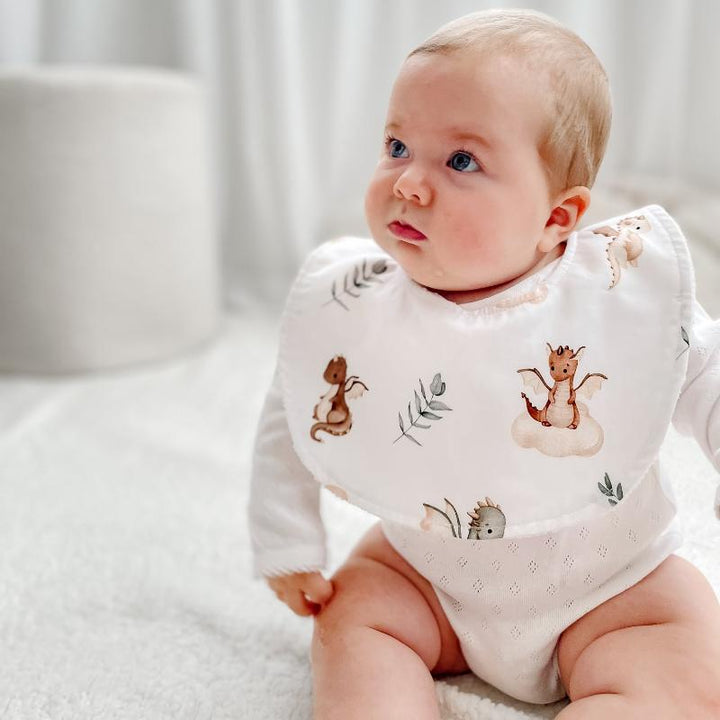 Baby sitting in a Mystique dragon print bib, crafted with organic cotton and absorbent microfiber to keep messes under control.