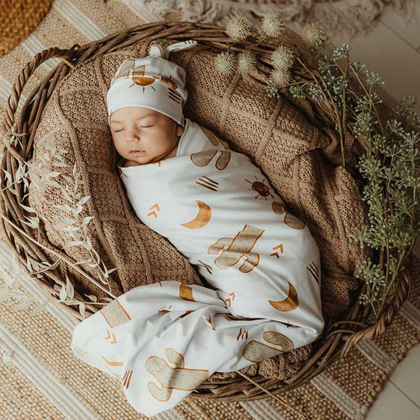 Baby sleeping in a basket covered in Snuggly Jacks Taupe Organic Knitted blanket  and wrapped in a stretch jersey swaddle with a knotted beanie