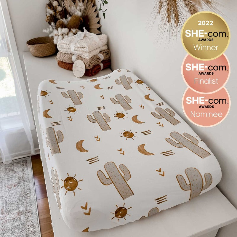 Change mat covered in an organic cotton bassinet sheet depicting a print of earthy cactuses, suns, moons on a white base in a setting with knitted blankets and australian native flowers in the background