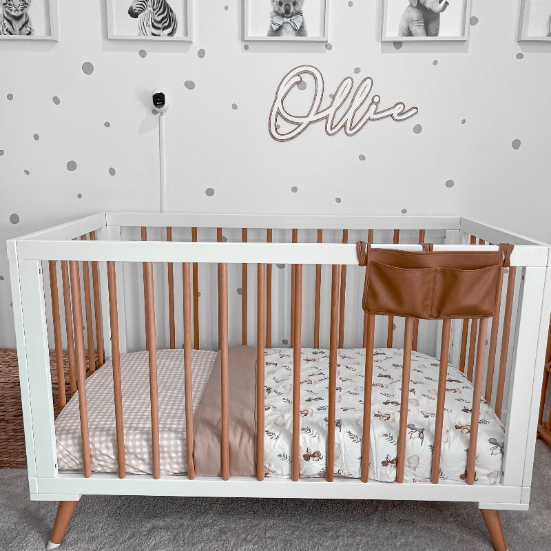 Pine cot with white accents set out in a nursery with Ollie written on the wall and cotton linen on the mattress.