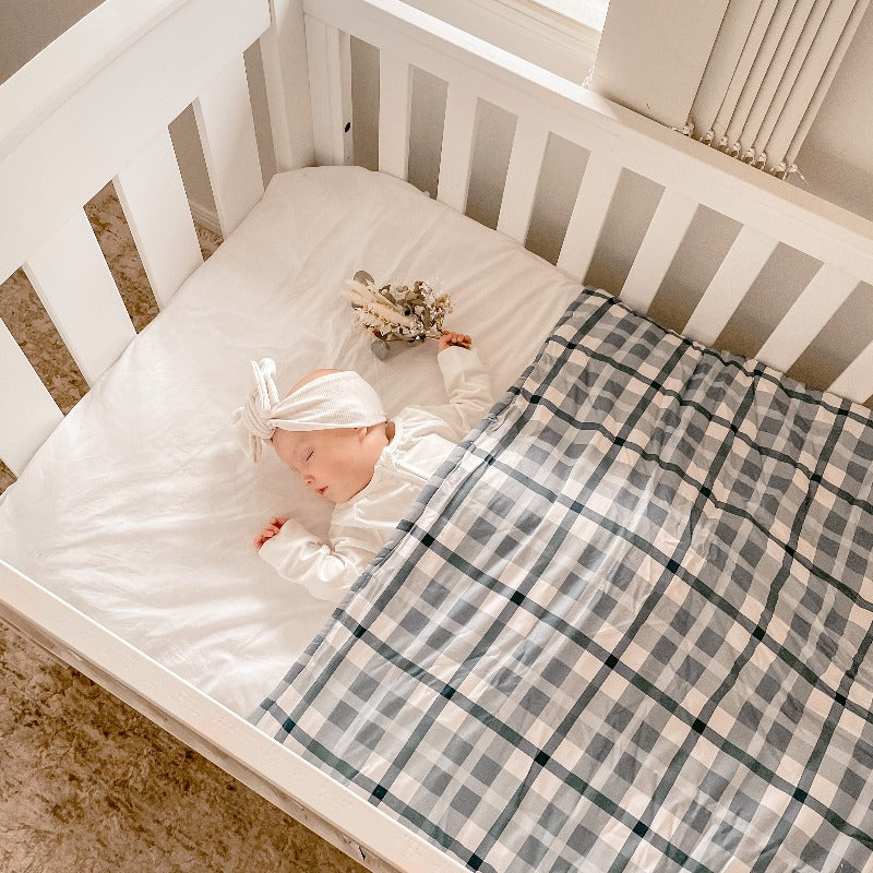 Baby wearing a white top knot laying in a white cot snug under a blue plaid cot quilt.