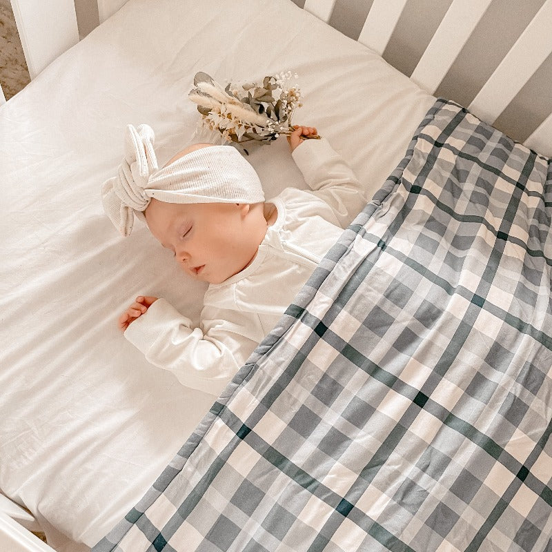Baby wearing a white top knot sleeping under a blue plaid cotton cot quilt