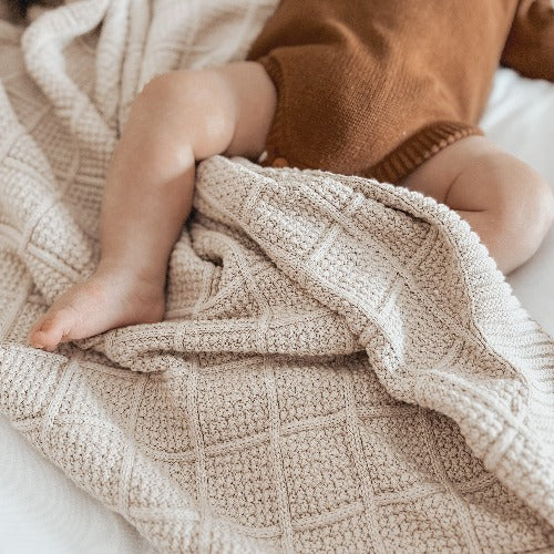 Baby laying on a bed with a brown onsie on and covered in a Snuggly Jacks Taupe Knitted Blanket