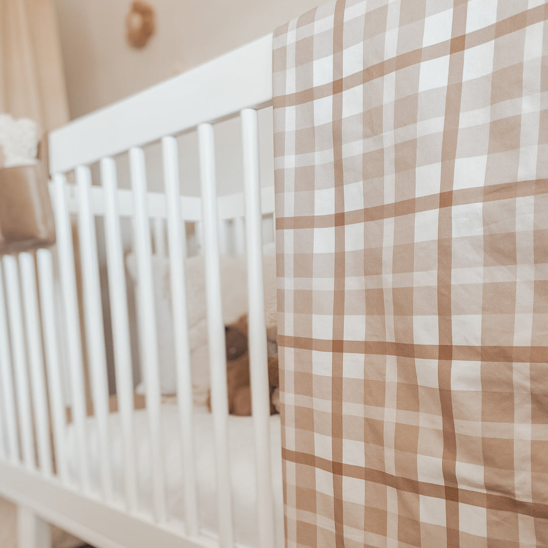 White pine cot with a brown plaid dimple dot minky blanket hanging over the side with a plush toy and pillows out of focus in the back ground