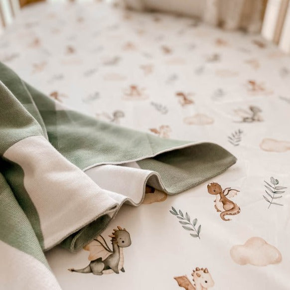 Pine cot made with snuggly jacks australia mystic fitted cot sheet with a green plaid knitted blanket draped over the setting.