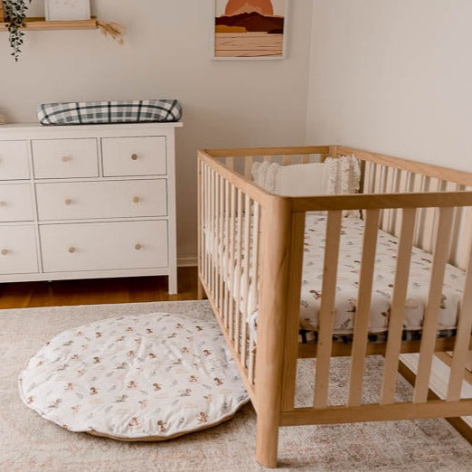 Wide shot of a nursery with soft colours featuring a pine cot, cotton play mat, change mat cover and a cotton fitted sheet all with the mystique dragon print on it.