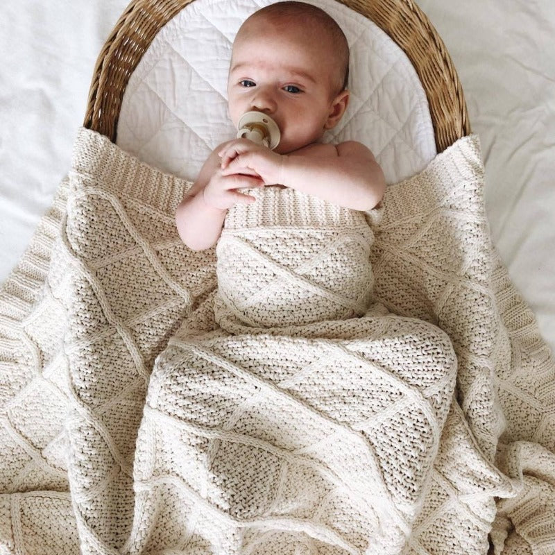 A cozy cream cotton knitted blanket by Snuggly Jacks, covering a sleeping baby in a bassinet