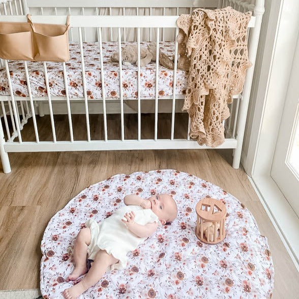 Nursery set out with a crocheted blanket hangint over the side of a cot with a plush bunny on the matress and a baby laying on a willow brown floral playmat on a wooden floor.