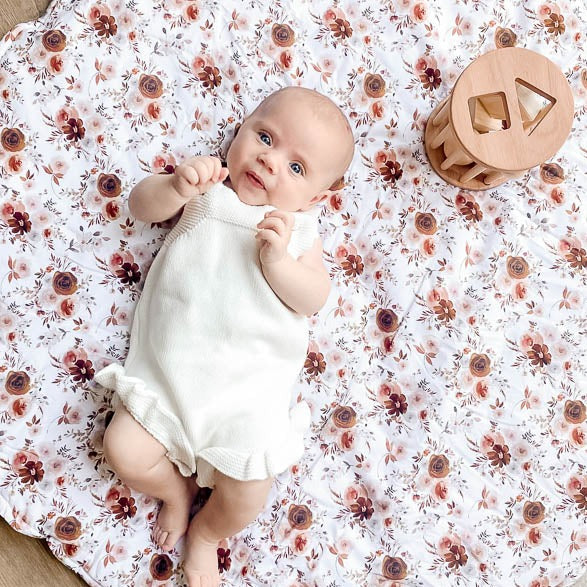 Over head photo of a blue eyed baby laying face up on a soft cotton play mat with a wooden childrens toy just by its head.