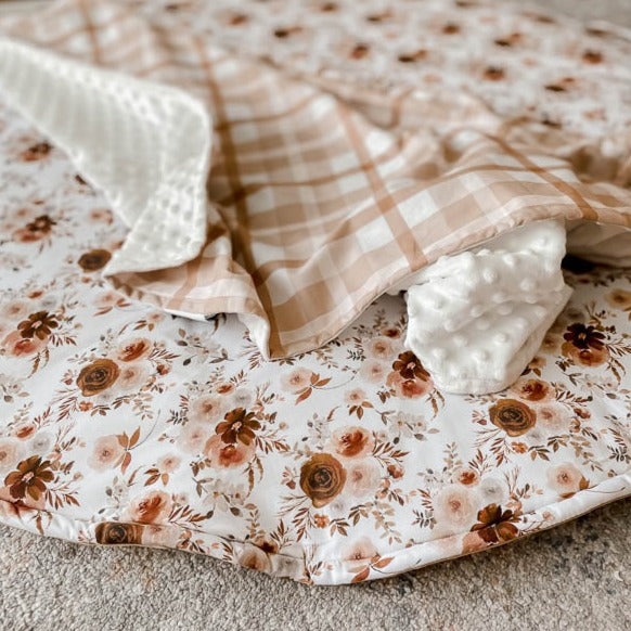 Close up shot of a play mat with flowers and leaves set out on it in a seemless pattern, there is a earthy plaid minky blanket laid across the play mat.